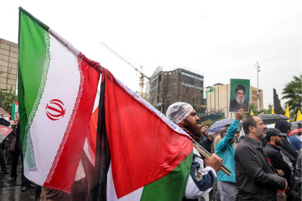 Um homem fica na chuva com uma bandeira iraniana e uma palestina durante um protesto anti-Israel na Praa Palestina, em Teer (Crdito: ATTA KENARE / AFP)